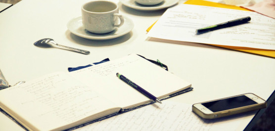 Auf einem Tisch liegen typische Utensilien für einen Workshop, wie zum Beispiel Stift, Notizbuch, Handy, Kaffeetasse.