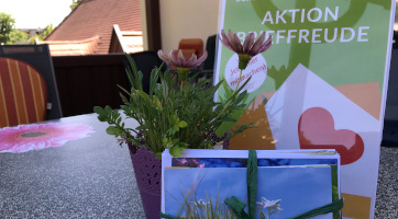 Ein Postkartenstapel liegt vor einem dekorativen Blumentopf auf einem Tisch, dahinter ein kleiner Tischaufsteller zur Aktion Brieffreude