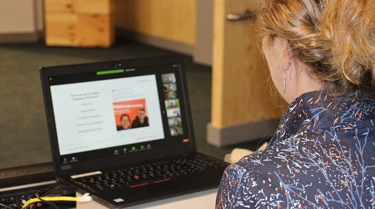 Foto: Eine Person sitzt vor einem Laptop, auf dessen Bildschirm unscharf in verschiedenen Kachel Personen und eine Präsentation zu erkennen sind.