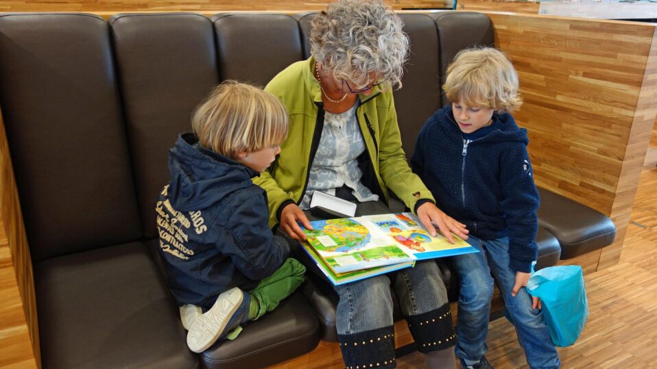 Eine ältere, grauhaarige Frau liest zwei Kindern aus einem Buch vor.