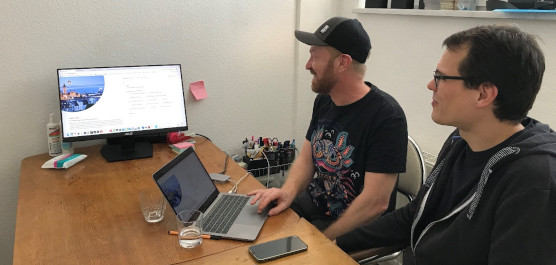 Zwei Personen sitzen vor einem Bildschirm und einem Laptop und arbeiten motiviert zusammen.