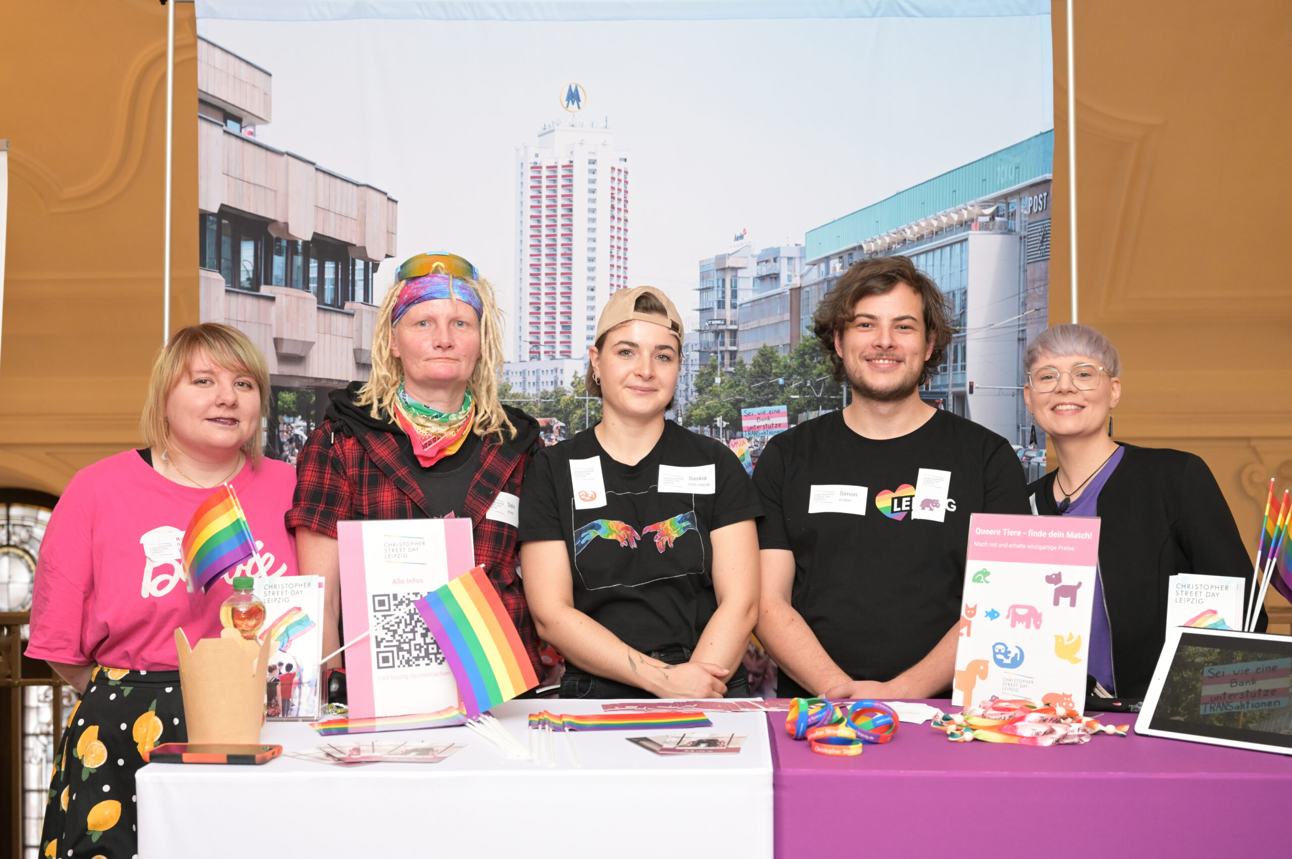 Fünf Personen am Stand des Christopher Street Day Leipzig. Fähnchen und diverse Kleidungsstücke in Regenbogen-Farben