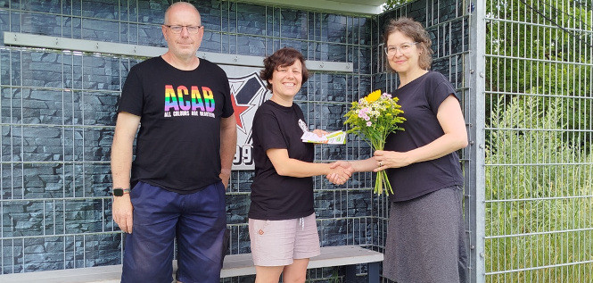 "Drei Personen vor dem Banner des Roten Stern Leipzig e.V. auf dem Rasenplatz, links im Bild die Mitarbeiterin der Freiwilligen-Agentur Leipzig mit einem Blumenstrauß als Dankeschön. 